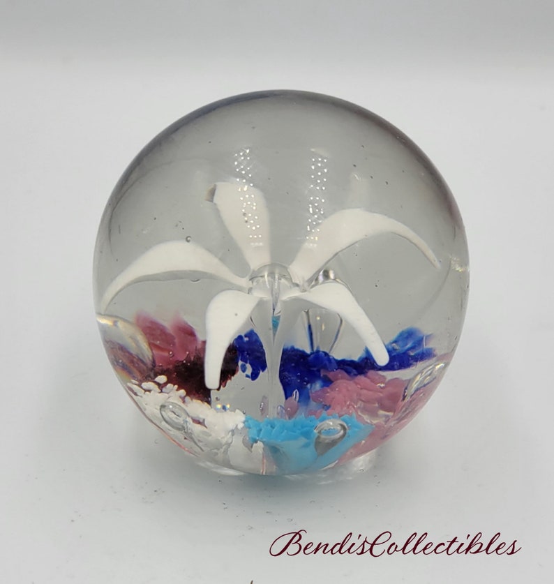 May include: A clear glass paperweight with a white flower inside. The flower has five petals and is surrounded by swirls of pink, blue, and turquoise glass.