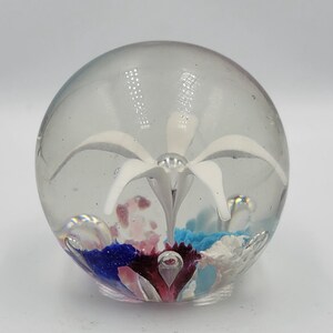 May include: A clear glass paperweight with a white flower and colorful glass accents inside.