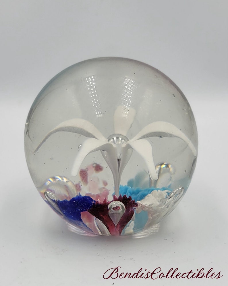 May include: A clear glass paperweight with a white flower and colorful glass accents inside.