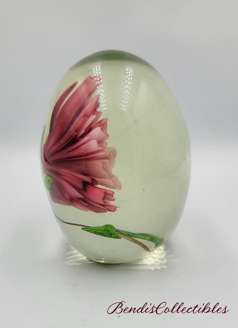 May include: A clear glass paperweight with a pink flower inside. The flower has green leaves and a stem.