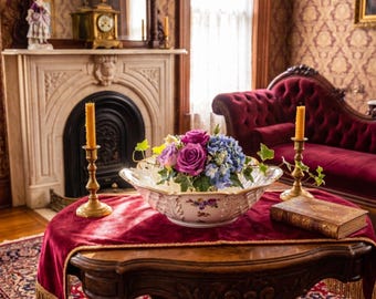 Puede incluir: Un cuenco de cerámica blanca con motivos florales contiene un ramo de rosas moradas, hortensias azules e hiedra verde. El cuenco se asienta sobre un camino de mesa de terciopelo rojo oscuro, flanqueado por dos candelabros de latón y un libro.