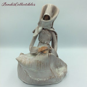 Puede incluir: Figurilla de cerámica blanca de una mujer con una capa con capucha sentada sobre una base texturizada.