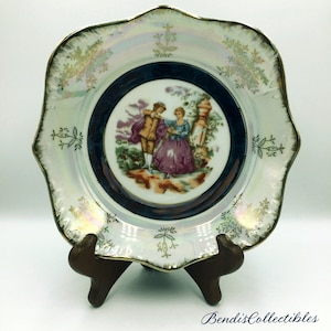 May include: A white porcelain plate with a scalloped edge and a gold rim. The plate features a central image of a couple in a garden setting, surrounded by a blue and gold border. The plate is on a wooden stand.