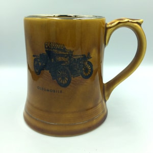 Puede incluir: Una taza de cerámica marrón con una imagen negra de un coche Oldsmobile antiguo. La taza tiene un borde negro y un asa marrón.