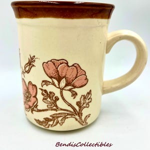 May include: Cream-colored ceramic mug with a brown rim and handle. The mug features a floral design in pink and brown. The text "BendisCollectibles" is printed at the bottom of the mug.