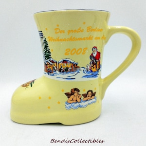 May include: A light yellow ceramic mug shaped like a boot, featuring a winter scene with Santa Claus and the text "Der große Berlin Weihnachtsmarkt an der 2008". The mug has a handle and is decorated with cherubs.