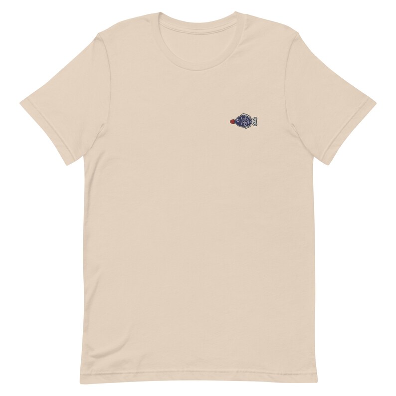 May include: A beige short-sleeved t-shirt with a small embroidered blue fish design on the front.