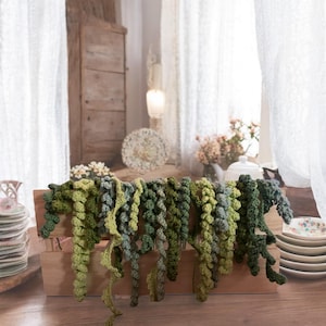 May include: A collection of green and blue crocheted garland strands hanging from a wooden shelf. The garland is made of small, round balls of yarn.
