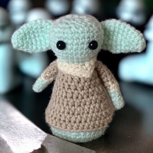 Puede incluir: Un juguete de peluche tejido a crochet que se asemeja a un personaje de la franquicia Star Wars. El juguete es verde y marrón con grandes ojos negros.