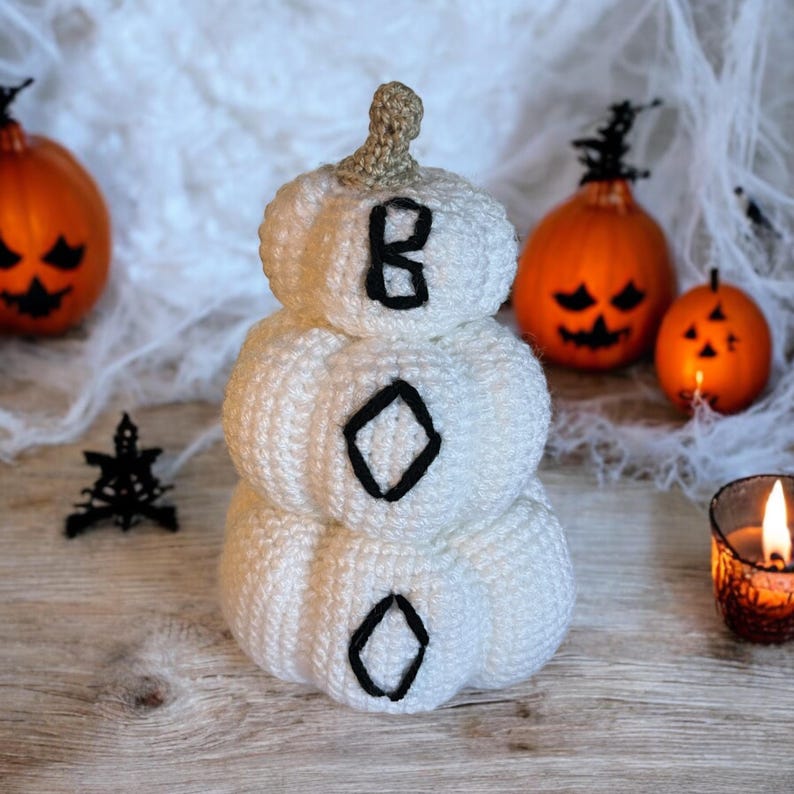 May include: A stacked white crochet pumpkin decoration featuring a tan stem and the word BOO embroidered in black yarn across the three tiers. The pumpkin is crafted with a textured yarn finish and sits upright as a singular decorative object.