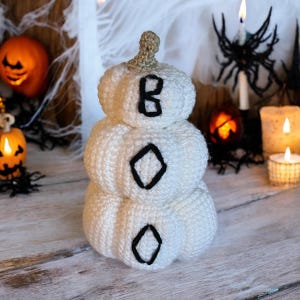 May include: A stacked white crochet pumpkin decoration featuring a tan stem and the word BOO embroidered in black thread across the three rounded segments. The object is crafted with a textured yarn finish and has a soft, rounded appearance.
