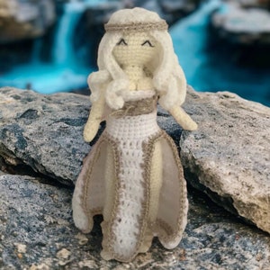 May include: A crocheted doll with long white hair, wearing a white dress with a gold trim, standing on a rock.