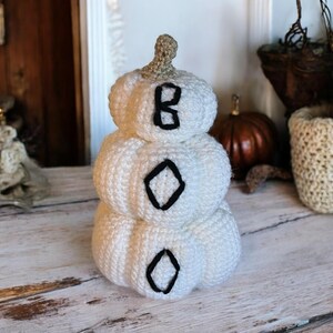 May include: A stacked white crochet pumpkin decoration featuring three tiers. Each tier is embroidered with a single black letter, spelling out the word BOO vertically from top to bottom. The pumpkin has a small, light brown crocheted stem.