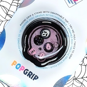 May include: A black PopGrip phone grip and stand with a Halloween theme. The interchangeable top features a pink skull design with a ghost, cross, and the word "RIP". The packaging includes the text "PopGrip".