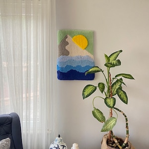 May include: A blue armchair with a throw pillow sits in front of a white wall with a colorful yarn art piece depicting a mountain and sun. A potted plant with green leaves and a brown burlap pot sits on a black table with a white ceramic vase and three white candle holders.