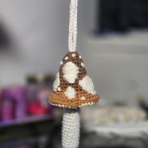 Op de afbeelding: Een handgemaakt gehaakt paddenstoelornament met een witte steel en een bruin en wit gevlekte kap. De paddenstoel wordt opgehangen door een wit gevlochten koord dat aan een zilverkleurige sluiting is bevestigd. De kap heeft een bruine rand.