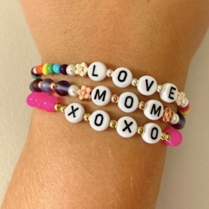 May include: Three colorful beaded bracelets with the words "LOVE", "MOM", and "XOXO" spelled out in white beads.
