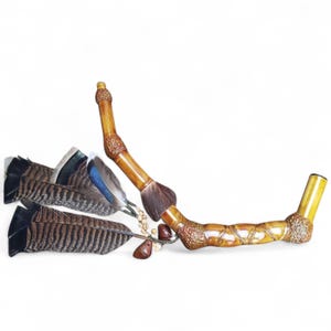 Pode incluir: Um cachimbo decorativo de bambu feito à mão com um design curvo. O cachimbo apresenta uma escova marrom, entalhes decorativos e é adornado com penas coloridas. O design inclui pequenas contas marrons e um pingente de madeira.