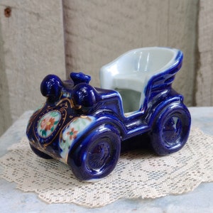 May include: A vintage blue ceramic car figurine with white interior. The car has gold accents and floral details. The figurine is sitting on a white lace doily.