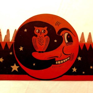 May include: A rectangular cardboard cutout featuring a red crescent moon with a smiling face and a black owl perched on its nose. The background is black with gold stars, and the top edge has a series of triangular points along the border.