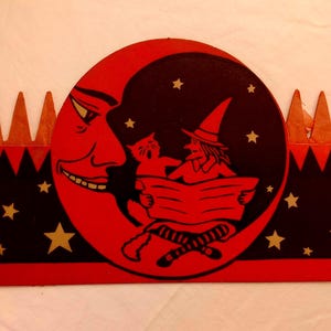 May include: A rectangular Halloween decoration featuring a large red crescent moon with a face, a black cat, and a witch reading a book. The design includes gold stars on a black background, a red border, and a row of orange triangular spikes on top.