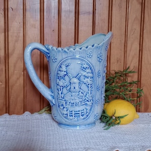 May include: A blue ceramic pitcher with a windmill design. The pitcher has a scalloped rim and a handle. It is sitting on a white crocheted doily.