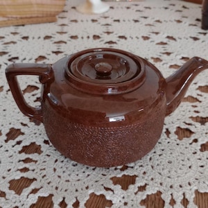 May include: A brown ceramic teapot with a textured lower half and a smooth, glossy lid. The teapot has a handle and a spout, and sits on a white lace doily.