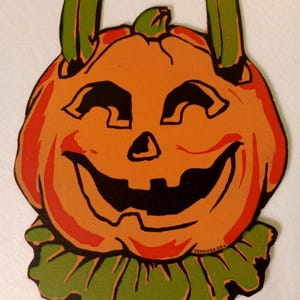 May include: A vintage orange paper pumpkin cutout with a black jack-o'-lantern face, green collar, and two green vertical tabs at the top. The text DENNISON U.S.A. is printed in small black letters on the lower right side of the pumpkin.