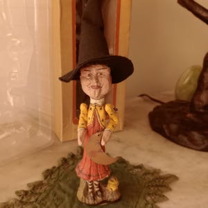 May include: A whimsical witch figurine wearing a black hat, yellow jacket, and red dress, holding a crescent moon. The figurine stands on a green lace doily. The witch is in a box.
