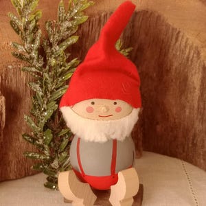 May include: A wooden gnome figurine with a red hat and suspenders. The gnome has a white beard and is sitting on a wooden base. The figurine is painted with a gray body and red accents. Decorative greenery is in the background.