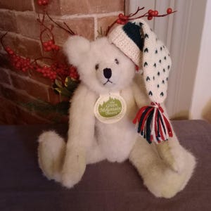 May include: White plush teddy bear wearing a white and green crocheted hat with red and white tassels. The bear has a tag that reads "The Green Mountain Bears".