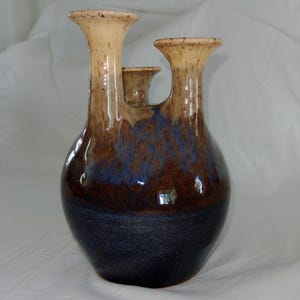 May include: A handcrafted ceramic vase with three spouts. The vase features a glossy finish with a color gradient, transitioning from a dark navy base to brown and tan at the top. The vase has a unique, organic shape.