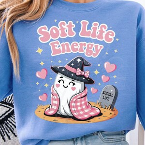 Può includere: Una felpa azzurra con la scritta "Soft Life Energy" in rosa. La grafica presenta un simpatico fantasma con un cappello da strega e avvolto in una coperta rosa e bianca. Una lapide recita "Social Life".