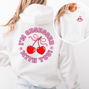 May include: White hoodie featuring a cherry design. The back of the hoodie has the text "I'M OBSESSED WITH YOU!" in pink, surrounding a pair of red cherries with a pink bow. The front has a smaller cherry graphic.