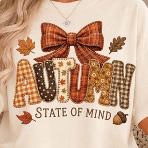 May include: A cream-colored t-shirt featuring a large graphic of a plaid bow above the word AUTUMN, where each letter has a unique pattern including gingham, polka dots, and leaves. Below, the text reads STATE OF MIND with an acorn and leaf.