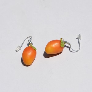 Op de afbeelding: Een paar mango-vormige oorbellen. Elke oorbel heeft een kleine, oranje en gele mango met een groene bladaccent. De oorbellen hebben zilverkleurige oorhaken en doorzichtige plastic oorbelruggen.