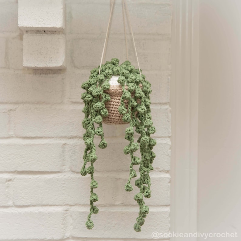 May include: A crocheted hanging plant with green leaves and a beige pot. The plant is hanging from a white brick wall.