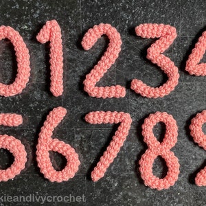 May include: Ten crocheted numbers, 0 through 9, in a light pink color, arranged in two rows on a dark gray textured surface. The numbers are made with a textured stitch and have a slightly puffy appearance. The text "@sookieandiycrochet" is visible at the bottom of the image.