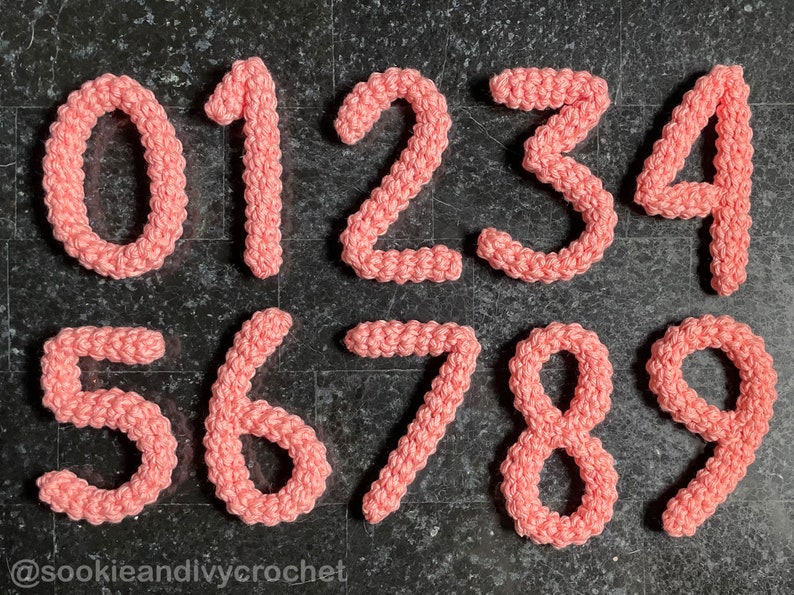 May include: Ten crocheted numbers, 0 through 9, in a light pink color, arranged in two rows on a dark gray textured surface. The numbers are made with a textured stitch and have a slightly puffy appearance. The text "@sookieandiycrochet" is visible at the bottom of the image.
