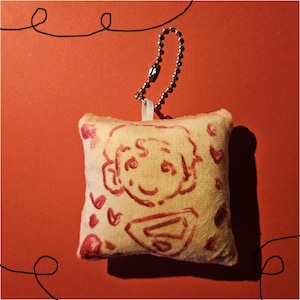 May include: A small, cream-coloured plush pillow featuring a red cartoon drawing of a person's face and hearts. It's attached to a silver ball chain. The background is a solid red.