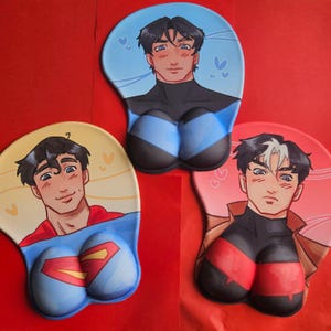 May include: Three ergonomic mouse pads with anime-style male character illustrations. One pad shows Superman, another a character in black and blue, and the third in red and black. The pads are designed for wrist support and are set against a red background.