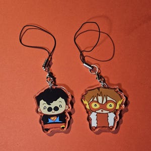 May include: Two acrylic charms featuring cartoon superheroes. One charm depicts a character with black hair, glasses, and a Superman-style outfit. The other charm shows a character with red hair and a Flash-inspired costume. Both charms have black cords.