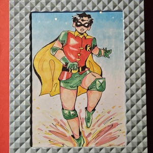 Könnte beinhalten: Gerahmte Illustration der Comicfigur Robin in einer dynamischen Pose. Das Kunstwerk zeigt Robin in seinem typischen roten, grünen und gelben Kostüm mit einem gelben Umhang. Der Rahmen ist hellgrau mit einem geometrischen Muster.