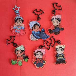 Puede incluir: Seis llaveros de acrílico con personajes estilo chibi que visten trajes de superhéroe y sombreros de cupcake. Los personajes llevan atuendos rojos, azules y negros con cierres en forma de murciélago o estrella en la parte superior de cada pieza.