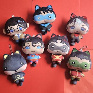 Puede incluir: Colección de peluches con diseños de superhéroes. Los peluches tienen orejas de gato y visten trajes icónicos. La paleta de colores incluye azul, rojo, amarillo y negro, sobre un fondo rojo.