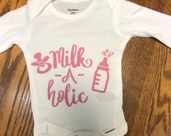 Può includere: Body bianco a maniche lunghe per neonato con testo rosa che recita "Milk-a-holic" e un'illustrazione rosa di un biberon e un ciuccio.