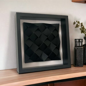 Può includere: Un'opera d'arte da parete incorniciata in nero con un design geometrico. Il design è realizzato in carta nera e piegato in un motivo tridimensionale.