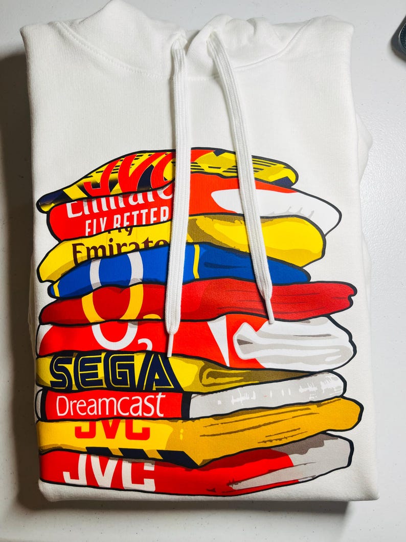 May include: White hoodie with a stack of colorful vintage video game and sports apparel logos printed on the front. The logos include Sega, Dreamcast, JVC, Fly Better, Emirates, and others.