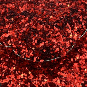 May include: Close-up of a pile of red glitter. The glitter is in the shape of small hexagons. The glitter is a bright, shiny red color. The glitter is in a small metal container.
