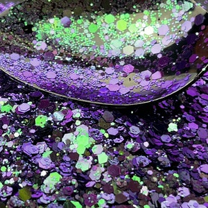 Puede incluir: Primer plano de una cuchara llena de purpurina morada y verde. La purpurina es una mezcla de formas hexagonales de varios tamaños. La cuchara refleja la purpurina, creando un efecto brillante. La purpurina está esparcida sobre una superficie oscura.